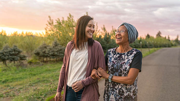 A woman walks arm in arm with her elderly mother on a sunset stroll.