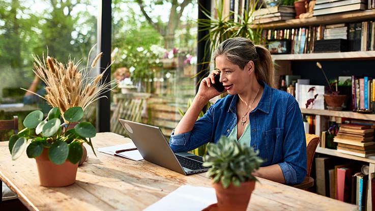 Sitting at a table in front of her laptop, a woman is speaking to someone on her smartphone