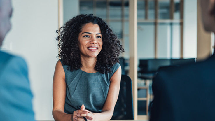 Sitting at a conference room table, a woman smiles confidently at a man and woman, both wearing suits and sitting across from her.