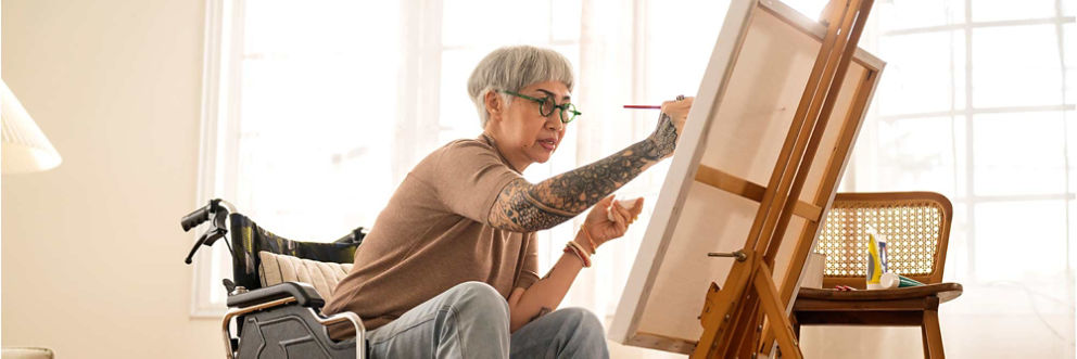 An older woman wearing glasses sits in her wheelchair while painting on a canvas.
