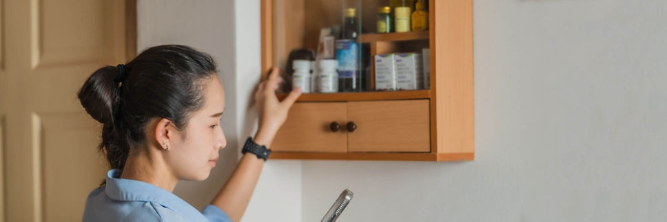 A woman glances at her phone while reaching into a medicine cabinet.