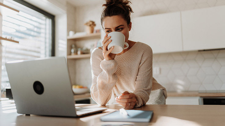 Sitting at her kitchen table, a young woman looks at her laptop’s screen while drinking a cup of coffee.