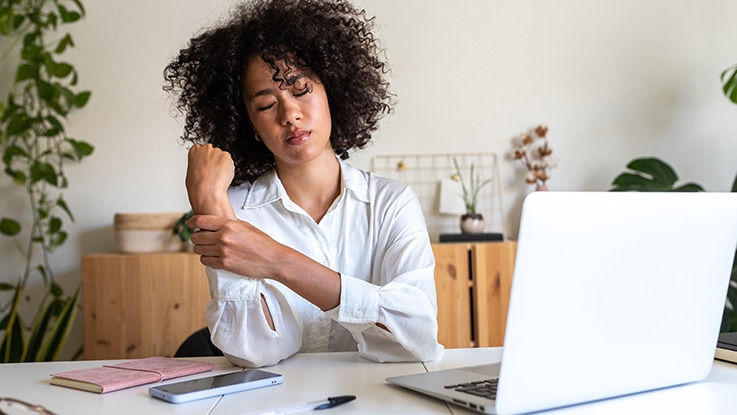 A woman works on her laptop in her home office. She's rubbing her wrist with a pained expression