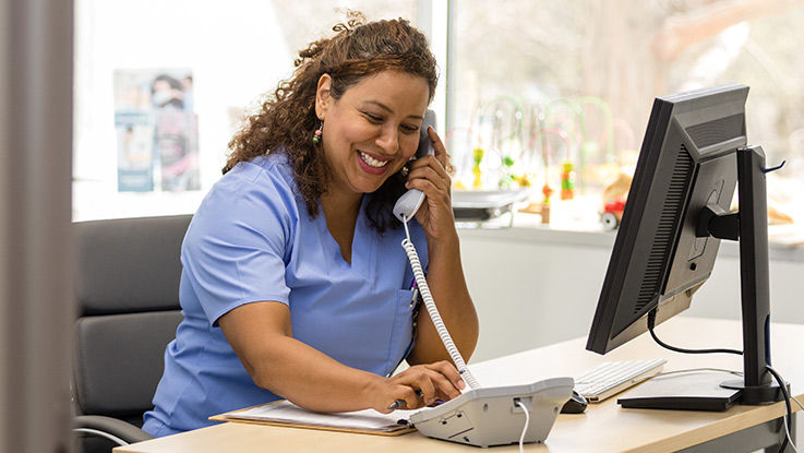 A nurse smiles as she sits in her office in front of a computer and speaks on the phone.