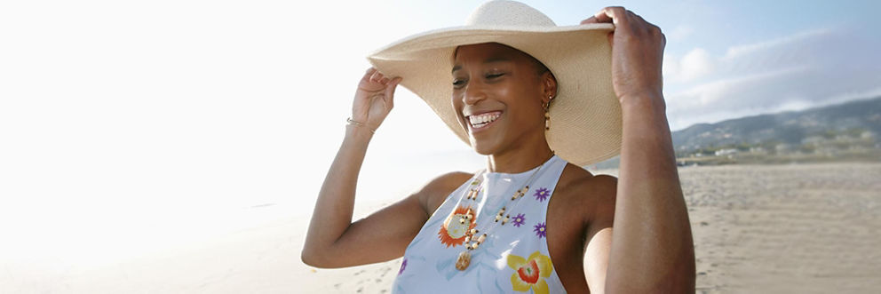 A smiling woman puts on a wide-brimmed straw hat at the beach.