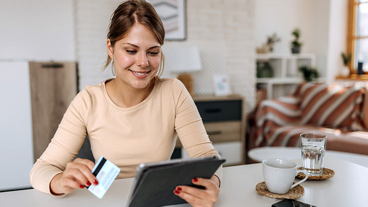 A smiling woman sits at a countertop holding a tablet device and a credit card.