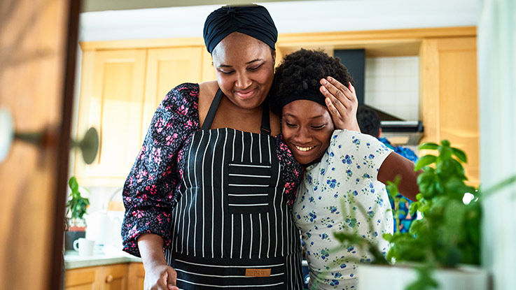 Standing in their kitchen, a smiling mother hugs her smiling daughter from the side while they both look at something in the distance