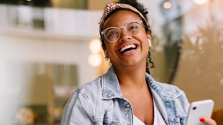 In a public café, a woman smiles widely as she listens to music and uses her phone.