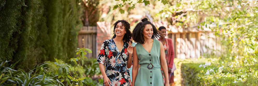 Two woman in dresses enter a sunny garden and smile as they look around.