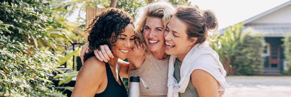 Three women wearing workout clothes laugh together outside.