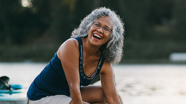 An older women wearing glasses and a navy blue tank top smiles while enjoying a nice day outdoors.
