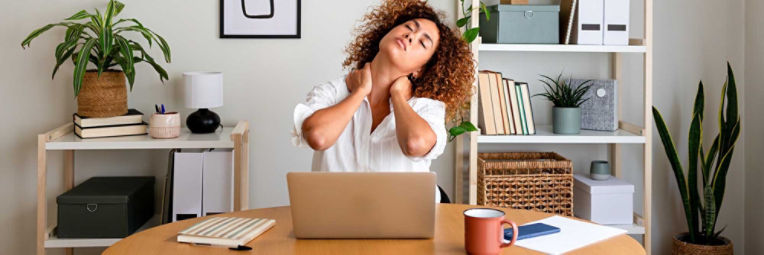A woman working on a laptop at a table puts both hands to her neck while stretching.