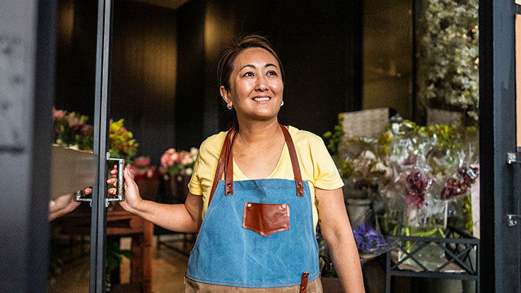 An owner of a flower shop stands in the front door of her business and looks out smiling.