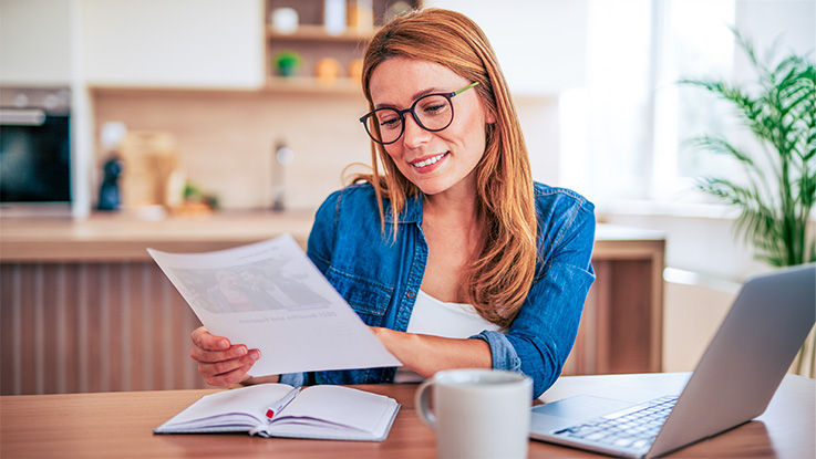 A woman sitting at her desk at home reads documents and information with her laptop open before her.