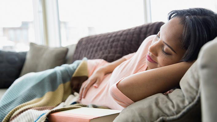 A pregnant woman naps on the couch with one hand on her belly.