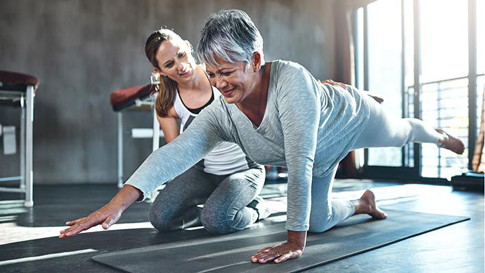 A smiling physical therapist rests her hand on a woman's back as she does a balance exercise.