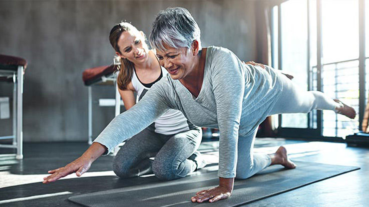An older woman does a complex yoga pose while her caregiver assists and smiles supportively.