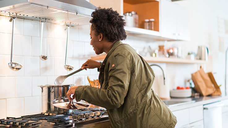 A woman cooks a meal in a soup pot on a stove in a bright, airy kitchen.