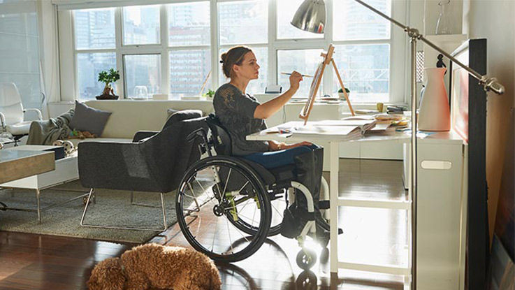 A woman in a wheelchair paints at a small easel on a desk.