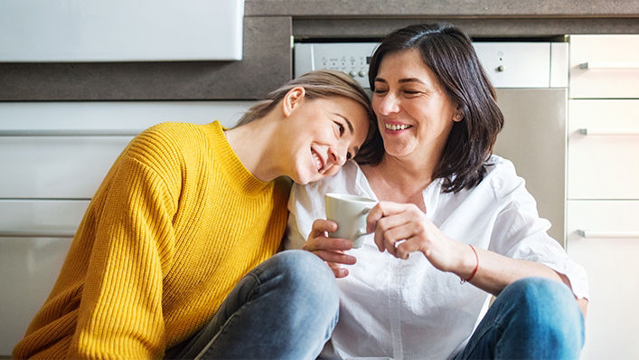 A daughter rests her head on her mother's shoulder while they laugh together.
