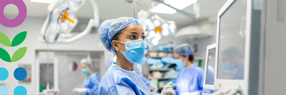 A CRNA wearing scrubs and a surgical mask checks a medical device in an operating room.