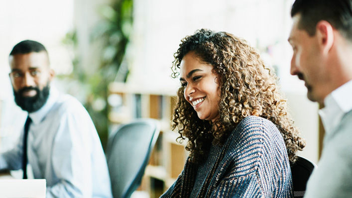 A woman seated at a conference table smiles and laughs as two colleagues share information with her