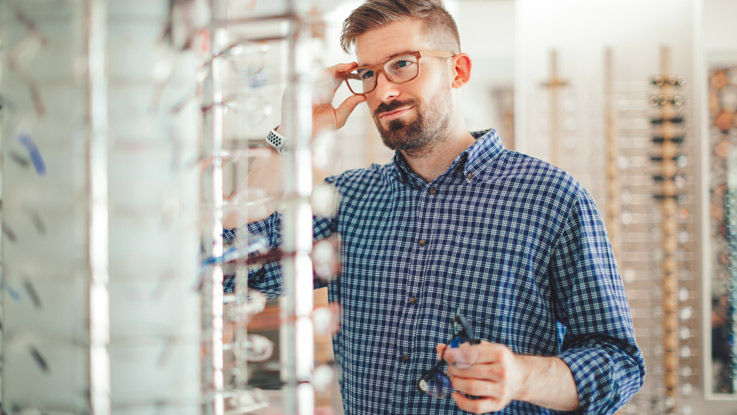 Man standing in an opticians shop trying on glasses.