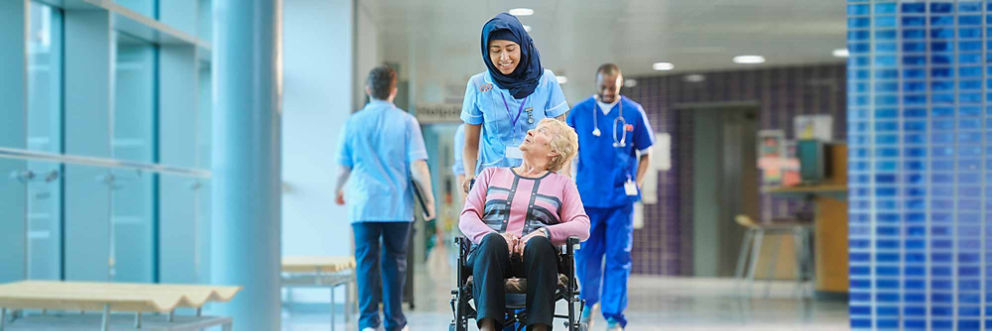 A woman volunteer pushes a man patient in a wheelchair down a hospital hallway.