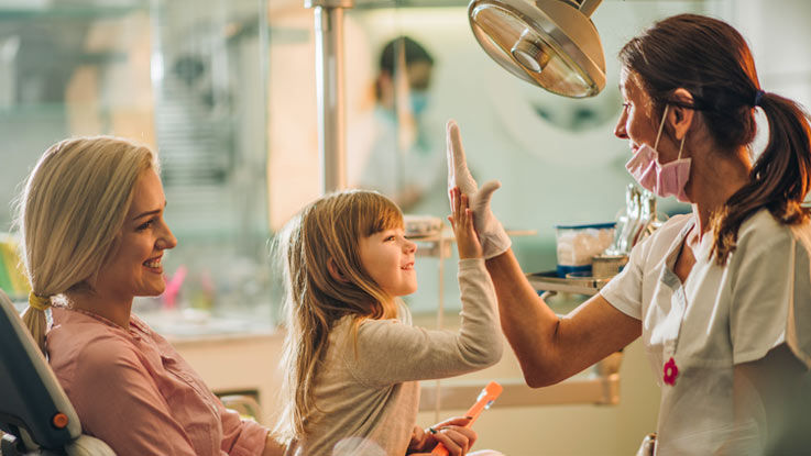 Sitting on her mother's lap in a dental exam room, a young girl high fives a dental assistant.