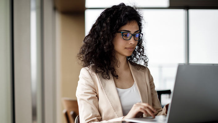 A woman wearing glasses and business attire works on her laptop