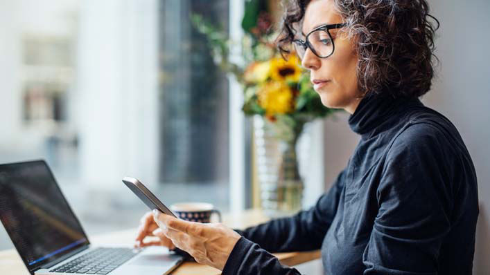 A woman wearing a black turtleneck and dark rimmed glasses glances down at her smartphone while working on her laptop.”