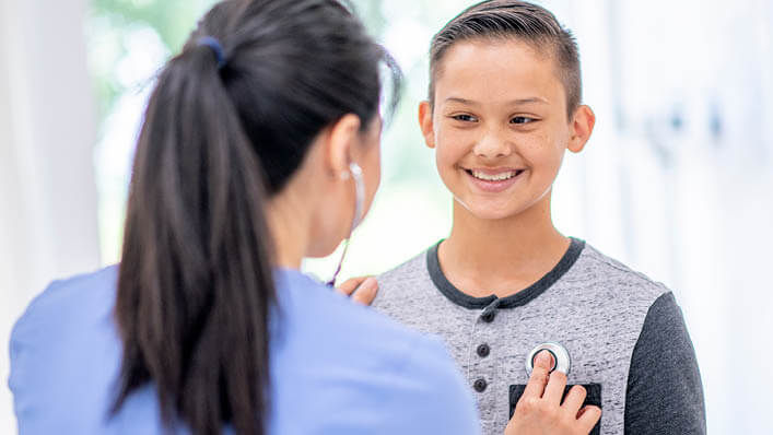 A young boy smiles as a doctor listens to his heart with her stethoscope.