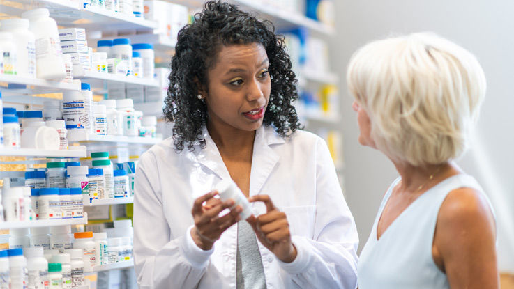 Pharmacist holding and pointing to a pill bottle, talking to an older woman in a pharmacy.