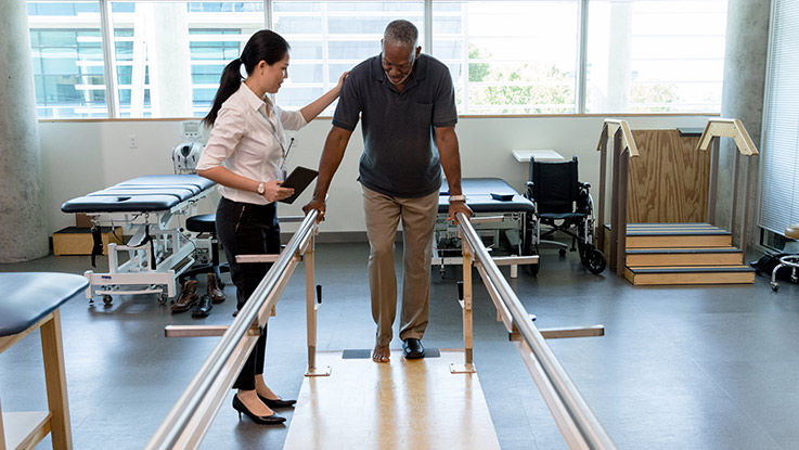 A physical therapist helps a middle-aged man position himself between parallel bars.
