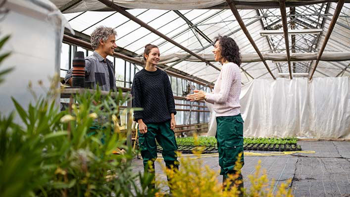 A group of three people stand talking in a circle in a greenhouse.