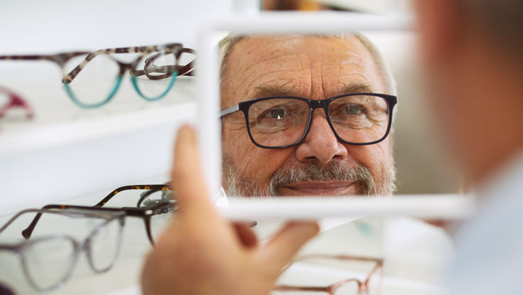 Man in an optical shop, trying on glasses and looking at himself in a mirror.