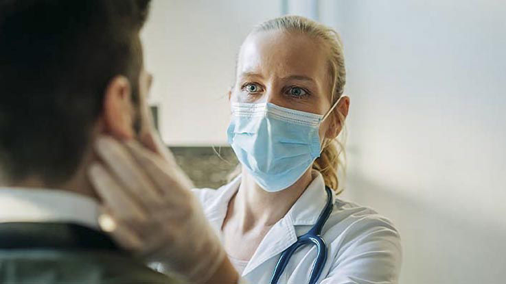 A female doctor wearing a face mask and gloves diagnoses a male patient by checking for throat swelling