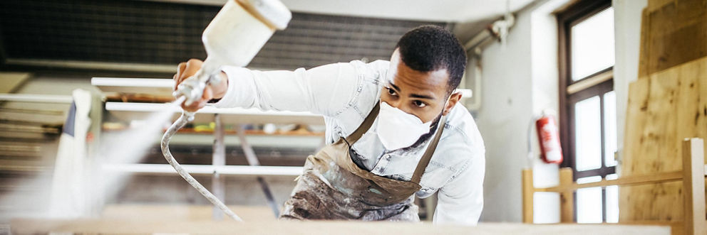 A young man has a strong grip on a power sprayer as he paints something in a workshop.