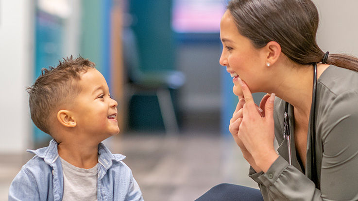 A pediatric therapist demonstrates a speech exercise to a child while he smiles.