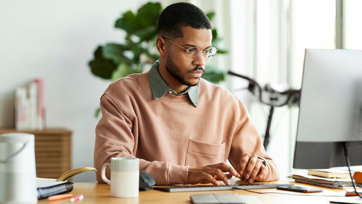 Man with glasses sitting at a desk and looking at a desktop computer.