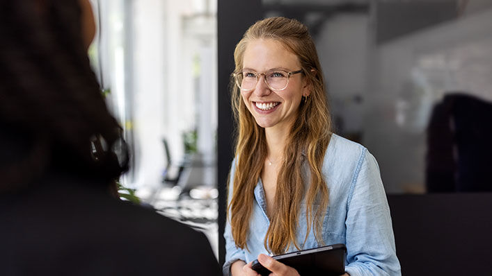 A woman wearing glasses and business attire smiles as she listens to a colleague