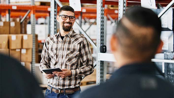 A man in a warehouse holding a tablet smiles as he speaks to other warehouse workers.