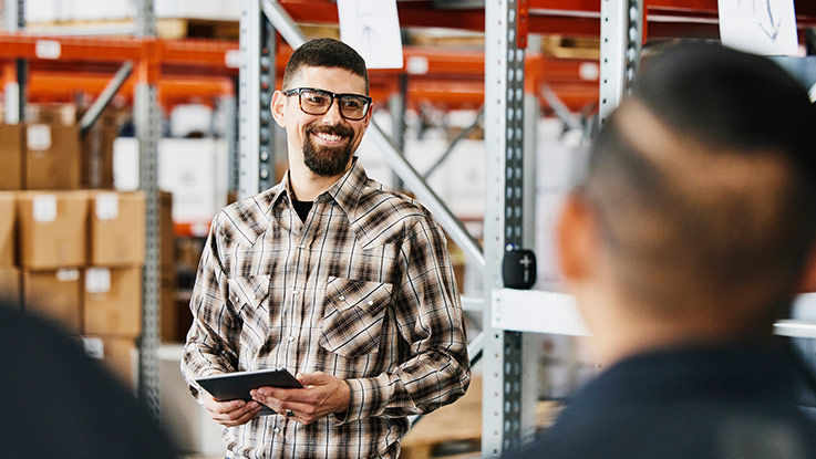 A man in a warehouse holding a tablet smiles as he speaks to other warehouse workers.