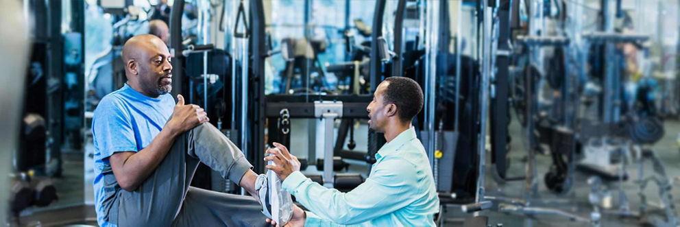 In a gym with various exercise equipment, a physical therapist helps his patient stretch his leg.