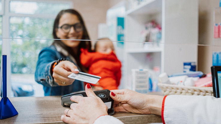 Cradling a baby in her arms at a pharmacy, a smiling young woman pays for her prescription using her credit card.