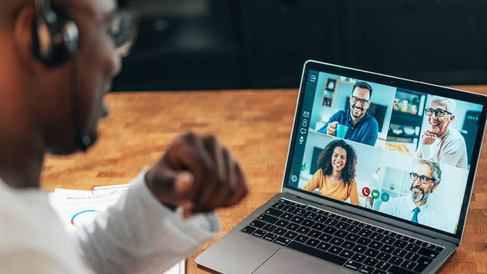 A man wearing glasses and headphones talks to four people on a virtual call using his computer