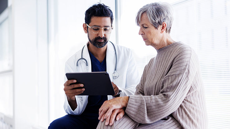 A doctor sits beside his patient and shows her information about her care on a tablet.