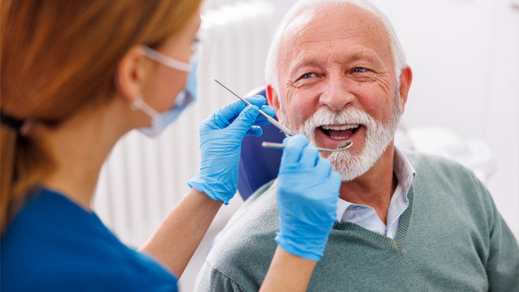 Sitting in an exam chair, a older man smiles before a dental assistant examines his teeth.