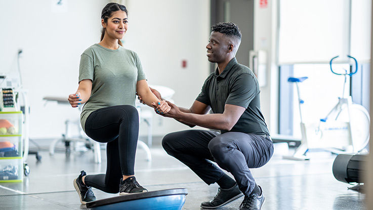  A woman lunges on a half ball to improve her balance while her physical therapist provides support.