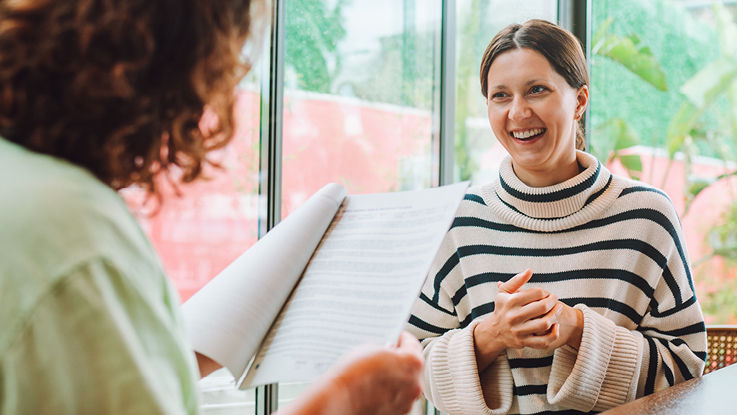 Two businesswomen sitting at a conference table smile and talk as they review the results of a report